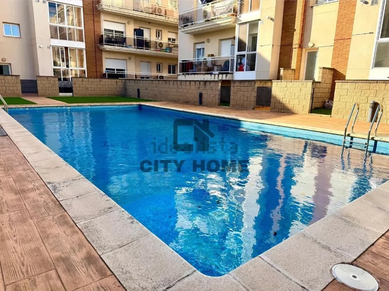 A bright blue rectangular swimming pool surrounded by concrete decking with multi-story apartment buildings in the background
