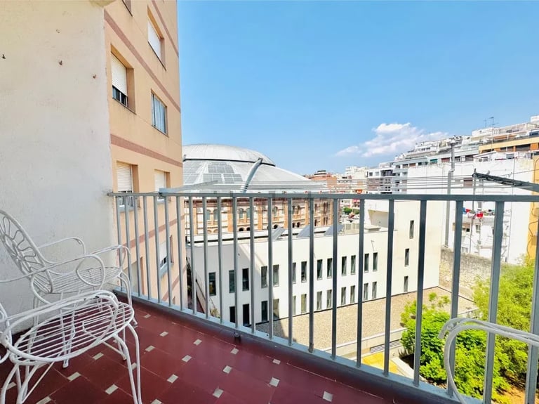 Urban apartment balcony with white chair overlooking red-tiled railing, city buildings, and clear blue sky