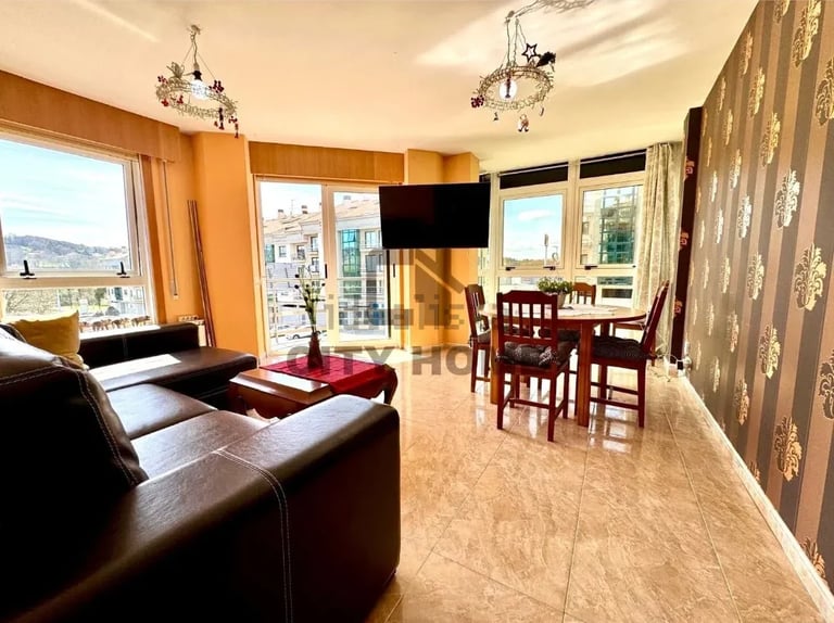 Modern open-plan living and dining room with leather sofa, TV, dining table, and large windows overlooking a coastal view