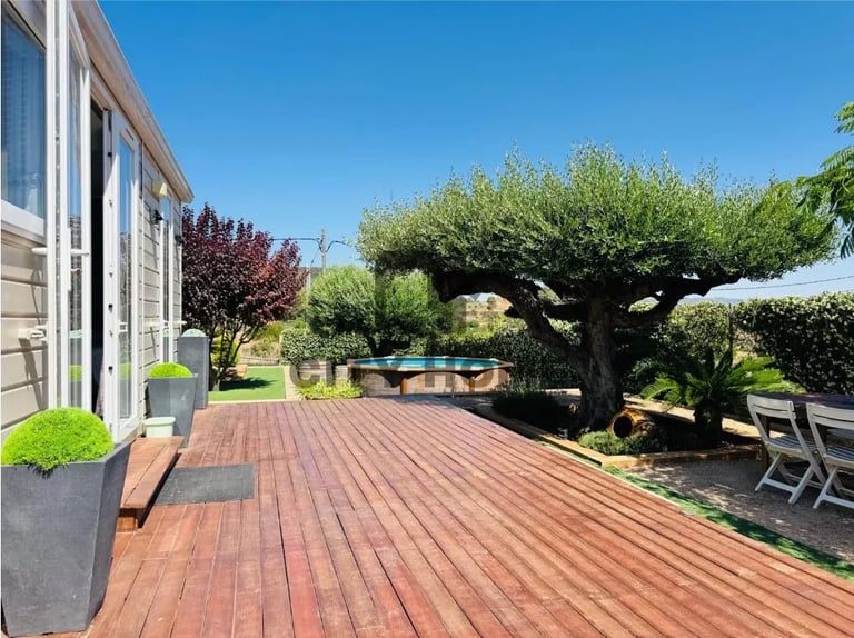 Residential backyard deck with reddish-brown wood flooring, mature shade trees, green lawn, and outdoor seating area under clear blue sky